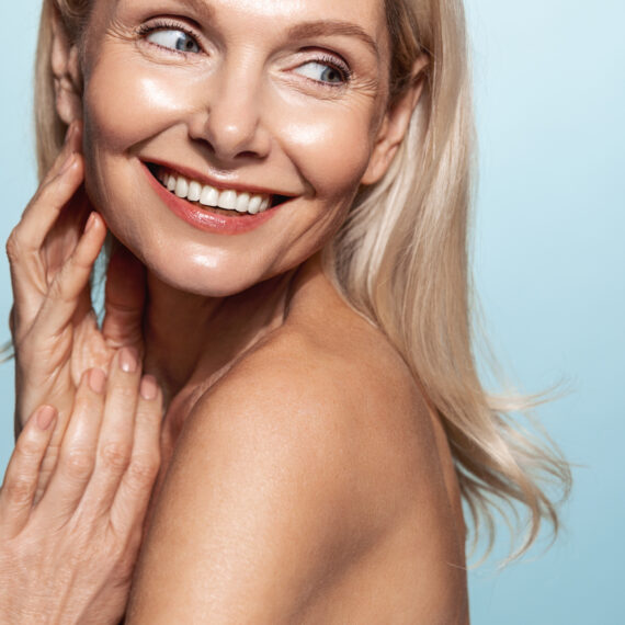 Happy middle aged woman with healthy, nourished glowing skin, smiling, looking behind her shoulder, blue background.