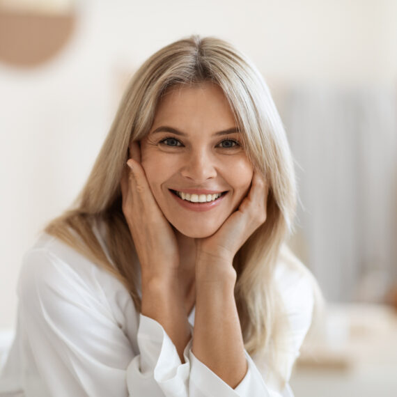 Beautiful blonde middle aged woman smiling at camera, touching her face, attractive lady doing morning face care routine, white bathroom interior, blurred background, closeup portrait, copy space