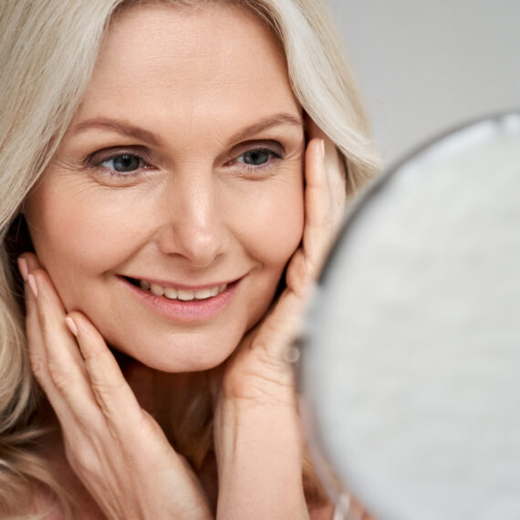 Happy 50s middle aged woman model touching face skin looking in mirror. Smiling mature older lady pampering, enjoying healthy skin care, aging beauty, skincare treatment cosmetic products concept.